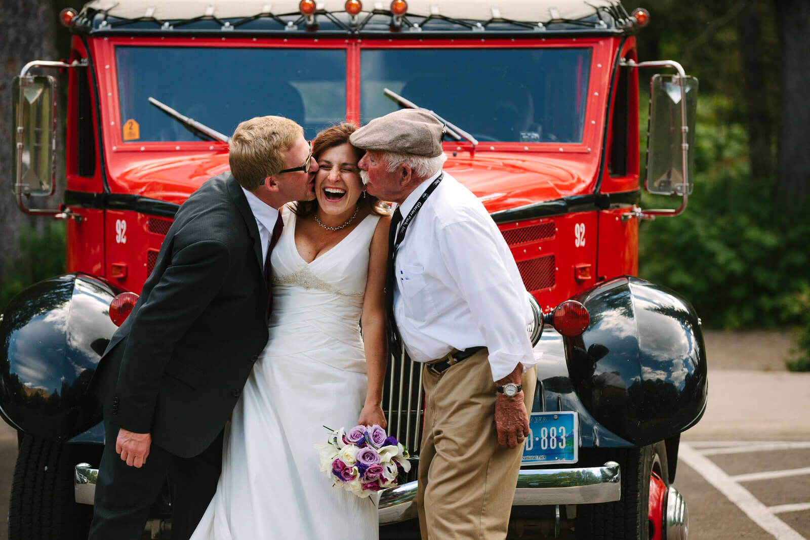 A bride laughs as she gets kissed by her new husband and the driver of a Glacier jammer bus during their Glacier National Park wedding in Montana