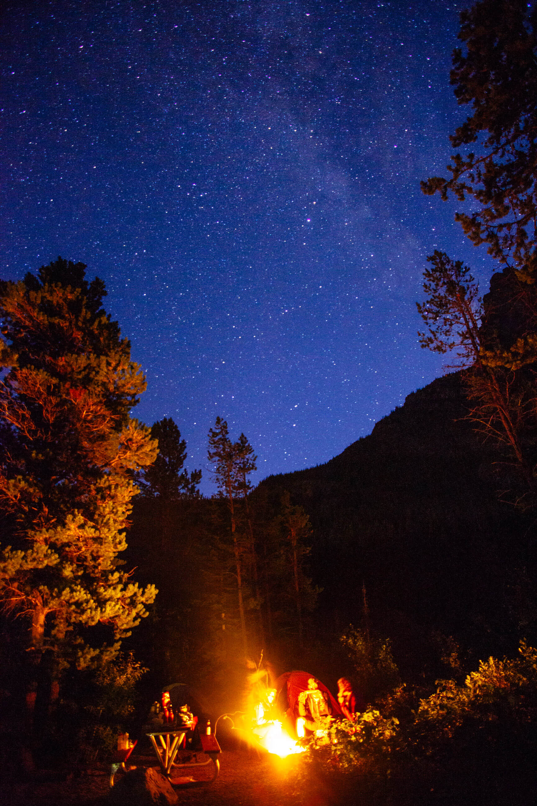 Campers sit near a glowing fire as stars populate the night sky in Glacier National Park in Montana
