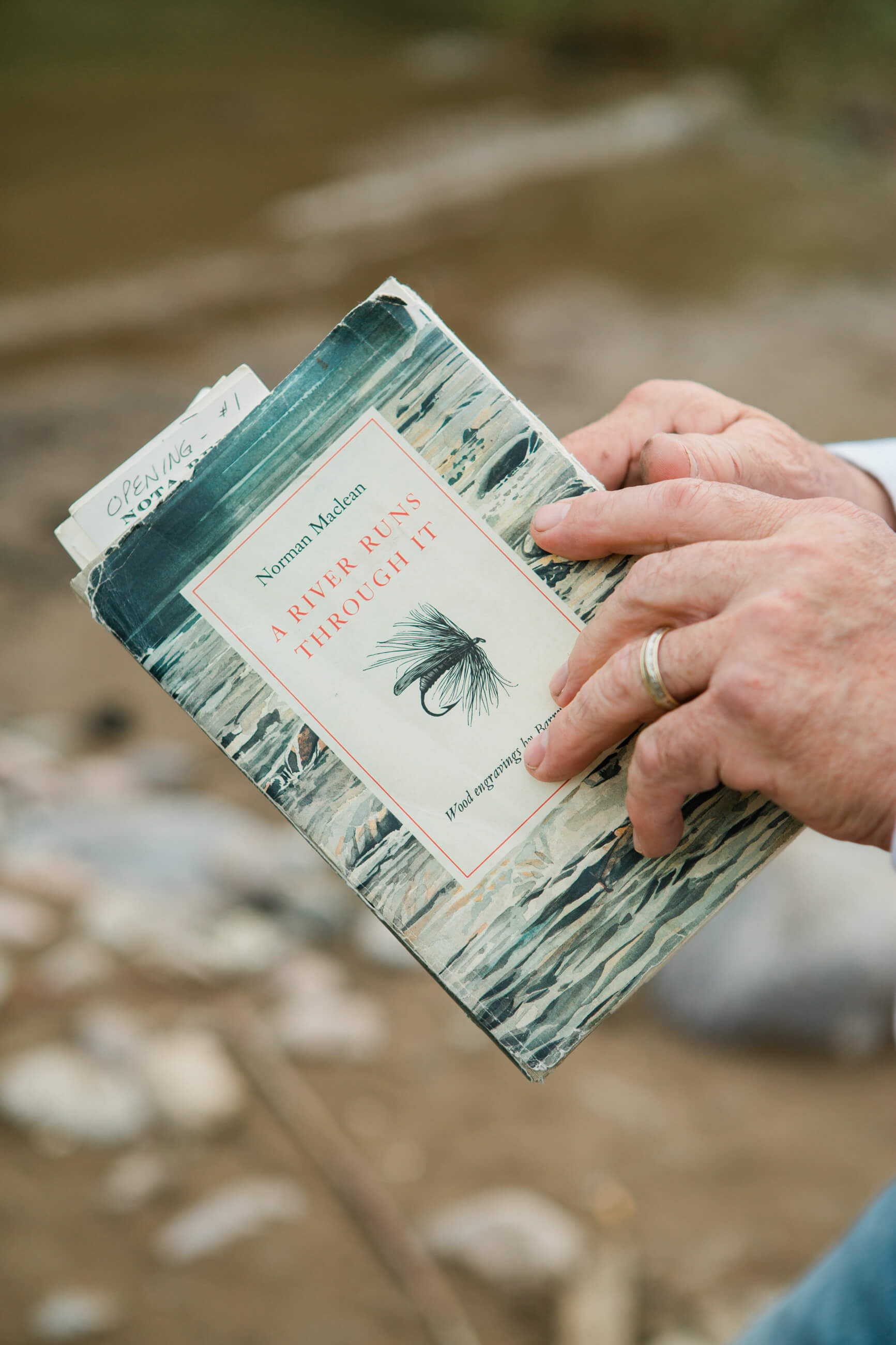 A man holds a worn copy of Norman Maclean's A River Runs Through it with the Blackfoot River in the background