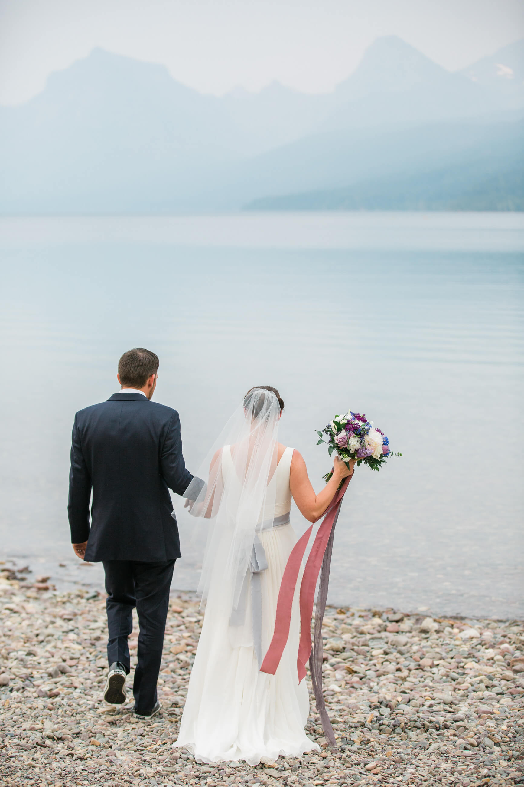 A bride and groom walk down a pebble beach to Lake McDonald in Glacier National Park during their Montana elopement