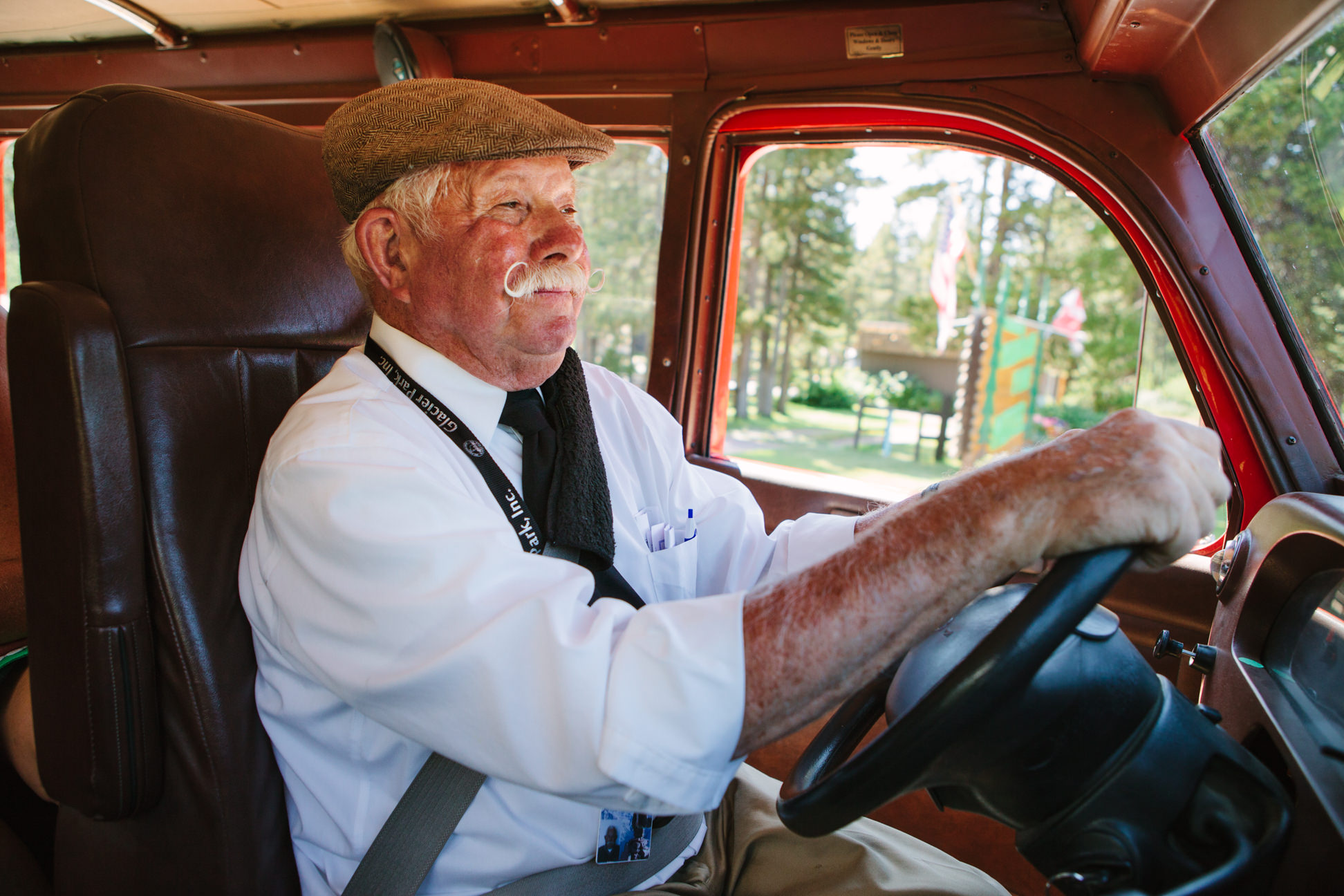 A red jammer bus driver with a handlebar mustache drives the Going-to-the-Sun Road in Glacier National Park