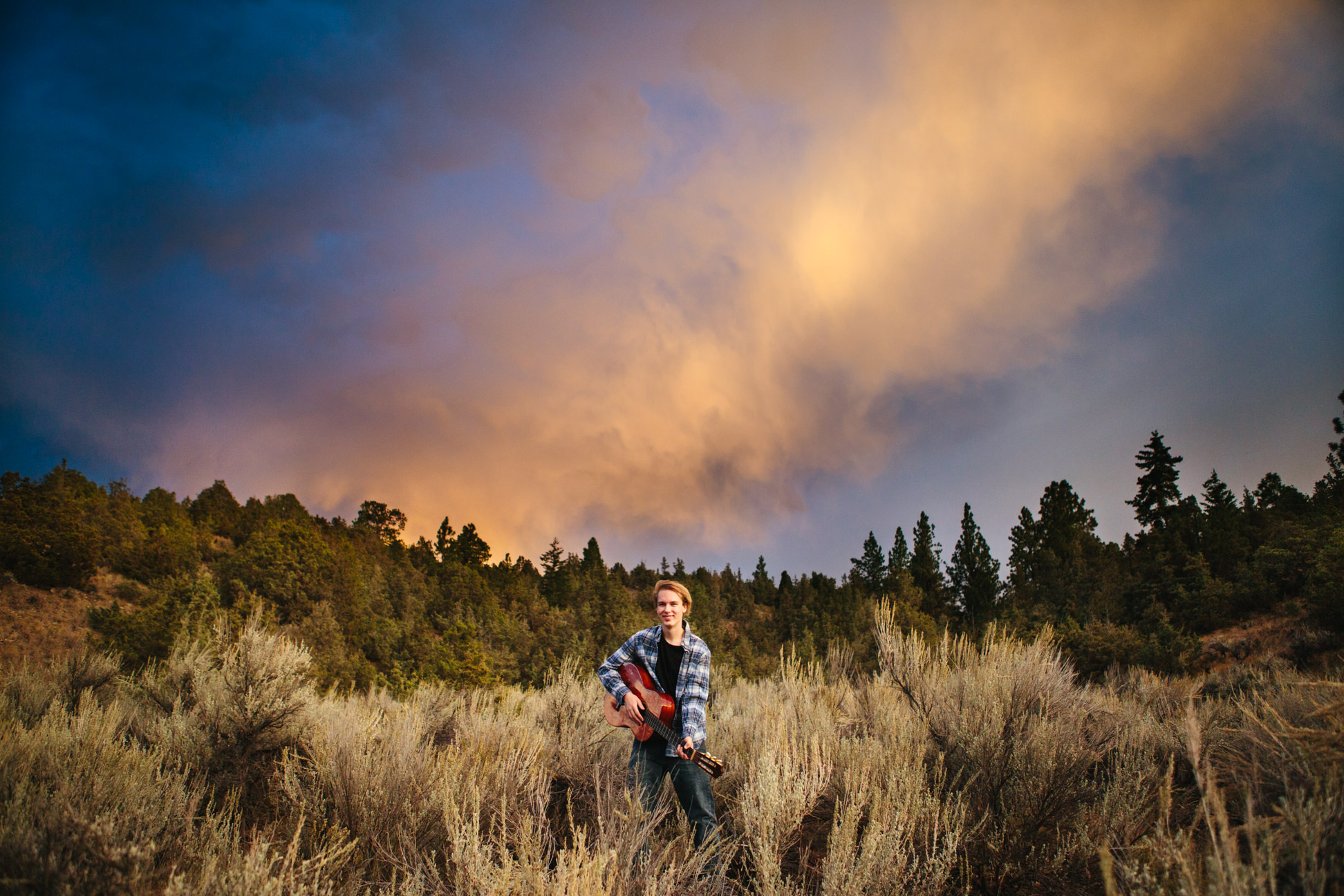 A young man stands with his guitar in a field of sagebrush at sunset in Corvallis Montana